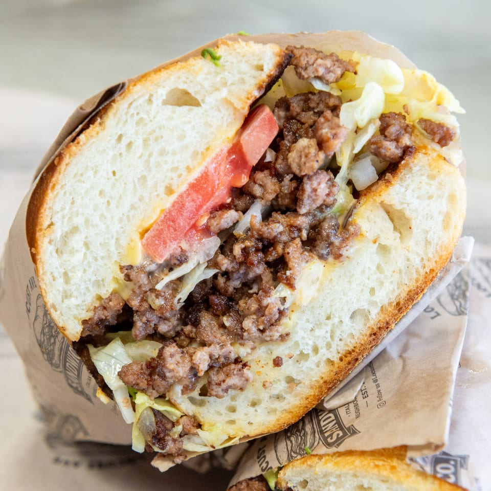 A close-up of a halved Sandy's sandwich, revealing ground meat, lettuce, tomato, and cheese.