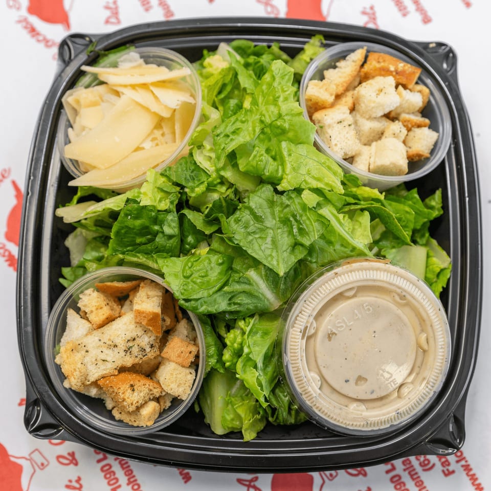 Caesar salad in a black to-go container with separate cups of croutons, Parmesan cheese, and dressing.