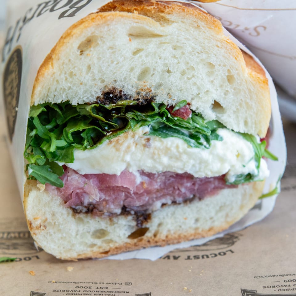 A close-up of a sandwich with meat, cheese, and greens on a baguette, wrapped in paper.